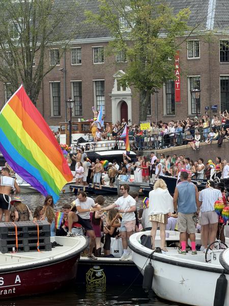 This vibrant photo captures a lively canal parade on the Amstel in Centrum, Amsterdam, filled with numerous people celebrating on boats and along the banks